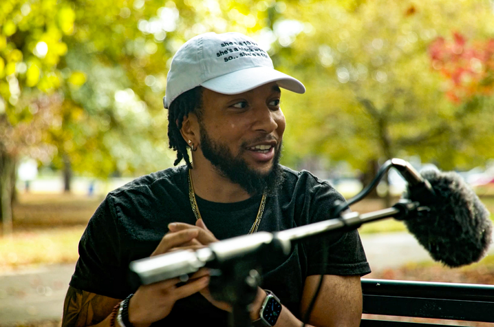 Student sitting on bench outside with boom microphone. He is wearing a white hat and black shirt, talking to someone to the side. 