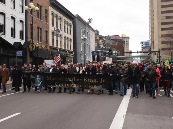 Large group marching in downtown Lexington streets with a banner reading Martin Luther King Jr. Day Celebration